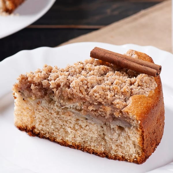 A slice of Brazilian apple crumble cake with cinnamon sticks on a white plate.