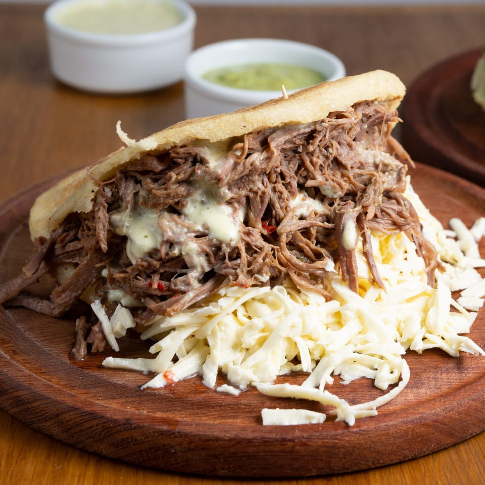 A Brazilian arepa with shredded beef, white sauce, and shredded cheese on a wooden board.