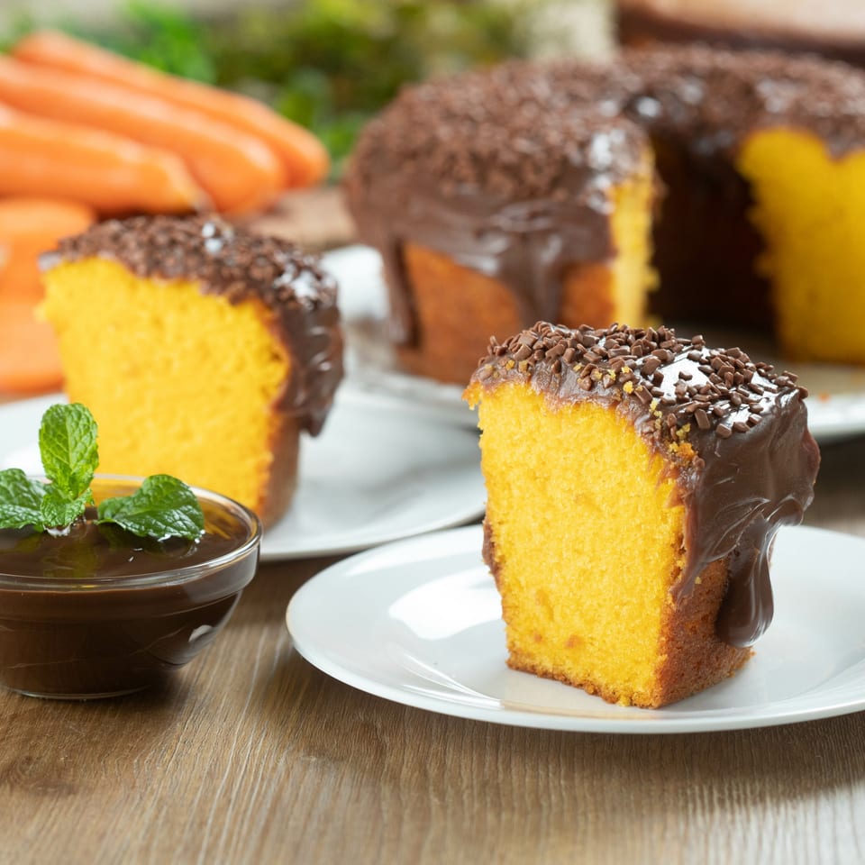 Slices of Brazilian carrot cake with chocolate frosting and sprinkles, with fresh carrots in the background.