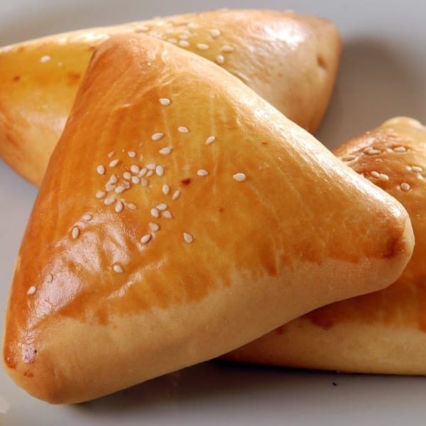 Three golden-brown Brazilian esfihas, triangular pastries topped with sesame seeds, on a white plate.