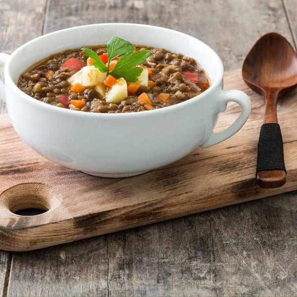 A white bowl of hearty lentil soup with carrots, potatoes, and parsley, next to a wooden spoon.