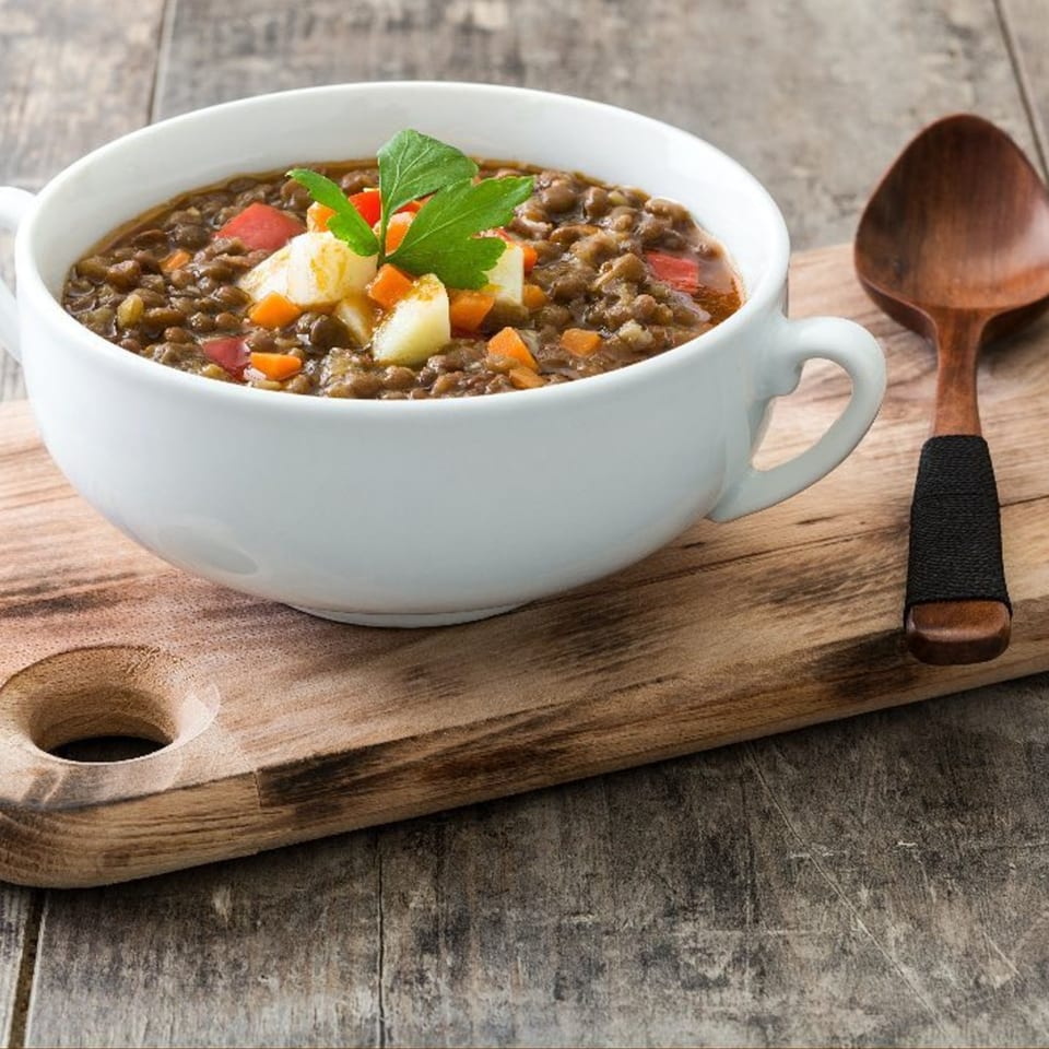 A white bowl of hearty lentil soup with carrots, potatoes, and parsley, next to a wooden spoon.