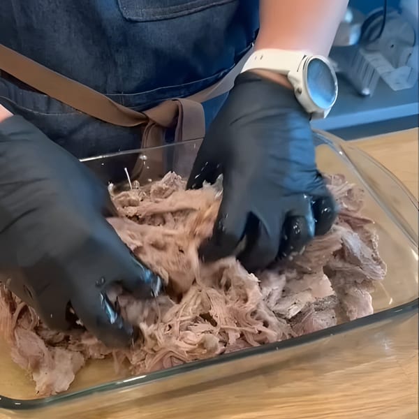 Gloved hands shredding cooked meat in a clear glass baking dish on a wooden surface.