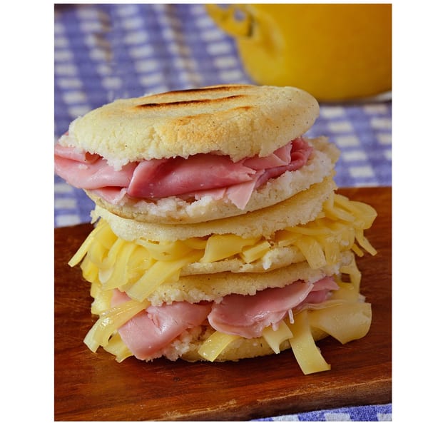Stacked Brazilian arepas with ham and cheese on a wooden board, with a blue checkered cloth.